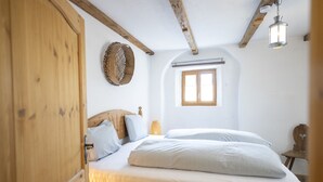 A bedroom with a wooden headboard, a bed with white linens, a woven wall hanging, a window with wooden shutters, and a hanging light fixture. - Chasa Rustica – Modern 3-Room Apartment with Traditional Charm in Sent (Sent)