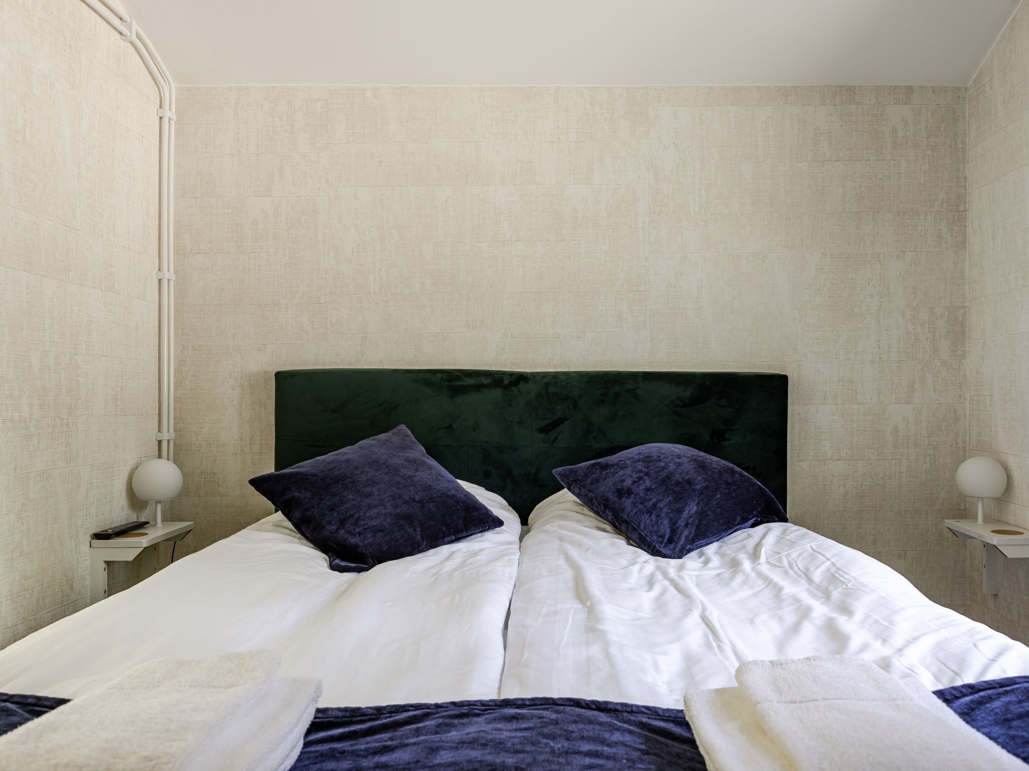 Una cama doble con ropa blanca y dos almohadas oscuras, un cabecero tapizado en verde y una pared beige lisa.