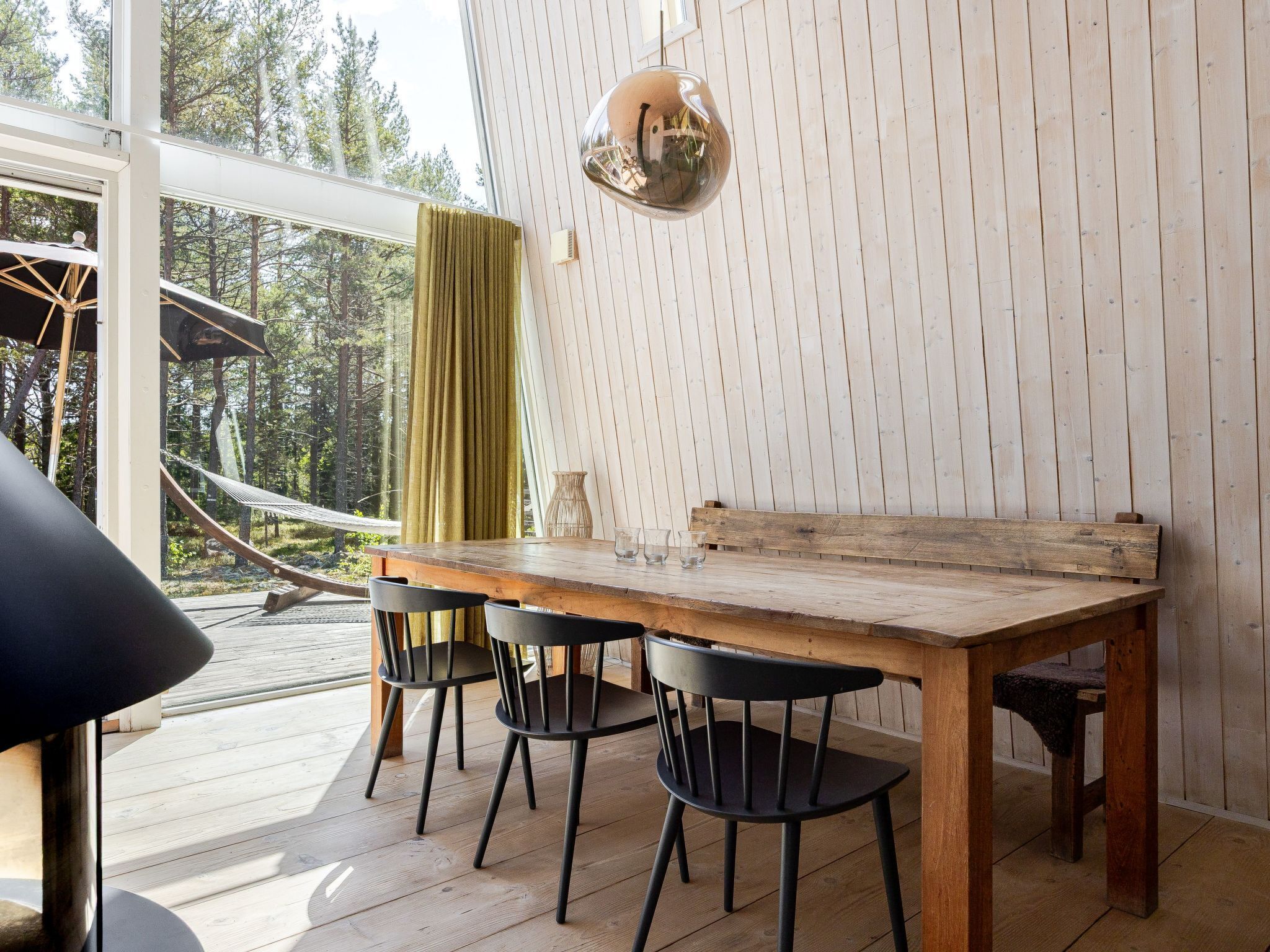 Een houten eettafel met vier stoelen, een hanglamp en uitzicht op een hangmat en bos door grote ramen.