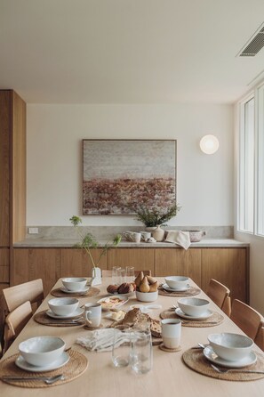 A dining area with a long table set for a meal, wooden cabinets, a large abstract painting on the wall, and a window providing natural light. - Osprey at Hawks Nest - Entire Property (Hawks Nest)