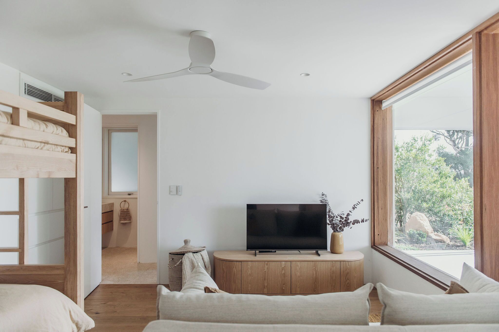 A modern bedroom with a TV on a wooden cabinet, a ceiling fan, and a large window with a view of greenery.