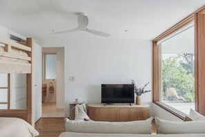 A modern bedroom with a TV on a wooden cabinet, a ceiling fan, and a large window with a view of greenery. - Osprey at Hawks Nest - Entire Property (Hawks Nest)