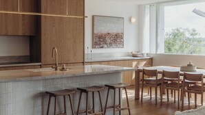 Eine moderne Küche mit Holzausstattung, einer großen Kochinsel mit Sitzgelegenheiten und einem Essbereich mit Blick auf Bäume durch das Fenster.