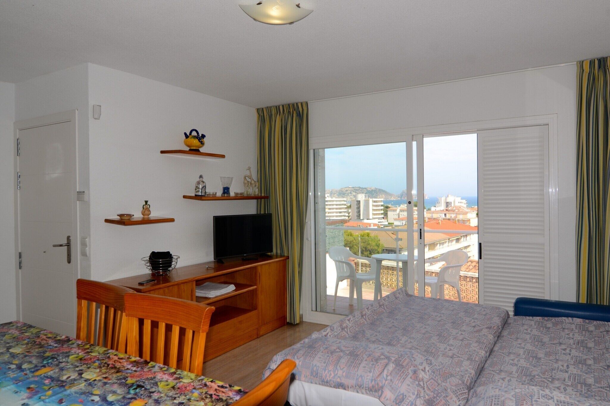 Et hotelværelse med en seng, et skrivebord med tv, en stol og en balkon med udsigt over byen.