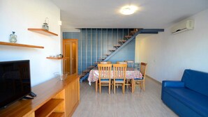A compact living space with a dining area, a TV on a wooden stand, a blue sofa, and a staircase leading to an upper level. - Apartment in Spain near Sandy Beach (Estartit)