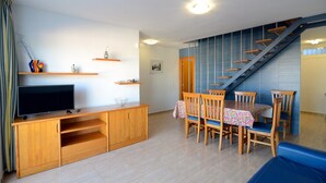 A compact living space with a TV on a wooden entertainment unit, a dining table with chairs, a staircase, and a blue wall. - Apartment in Spain near Sandy Beach (Estartit)