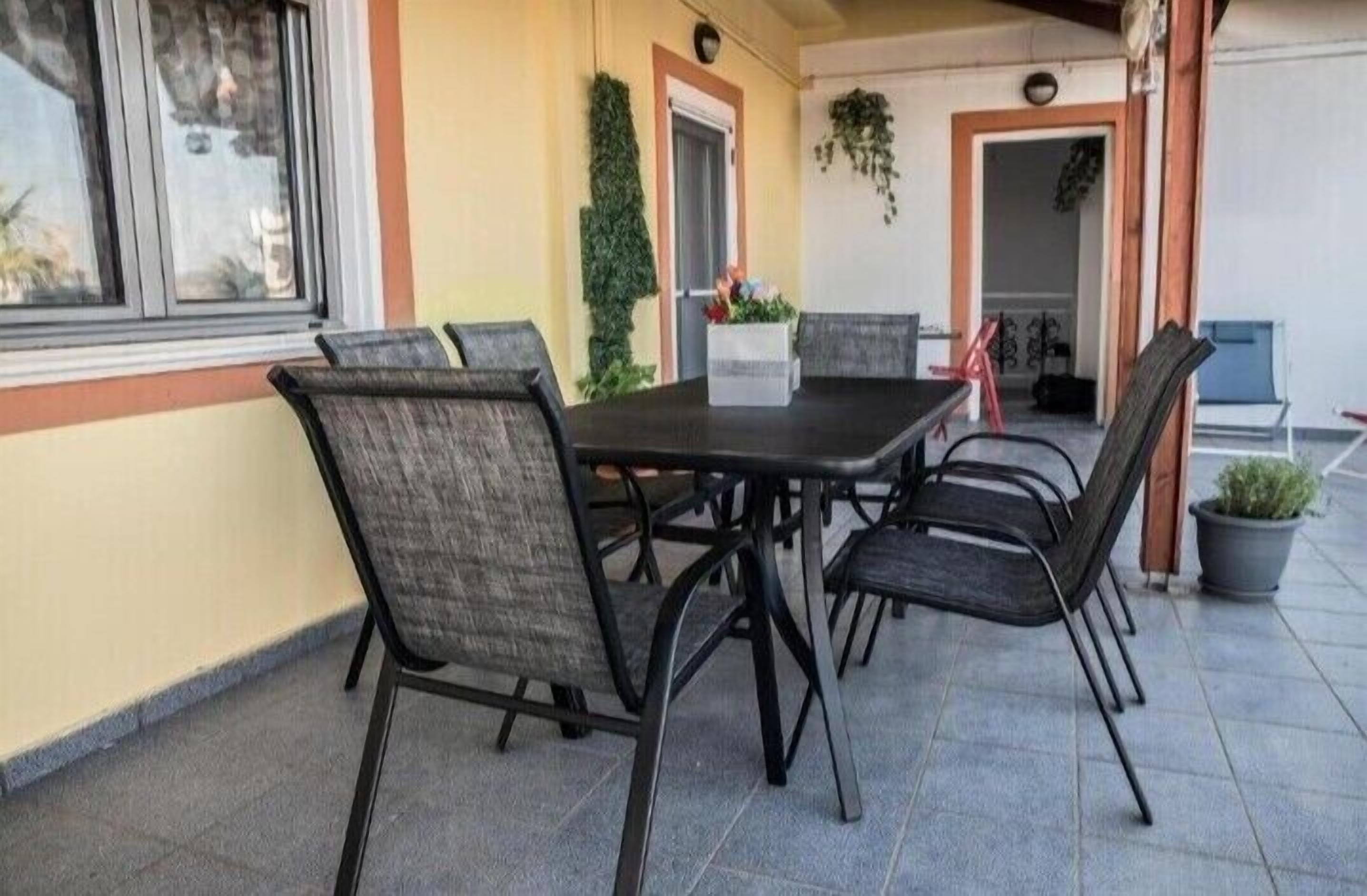 Une terrasse couverte avec une table et des chaises, une plante en pot et une porte donnant sur une autre pièce.