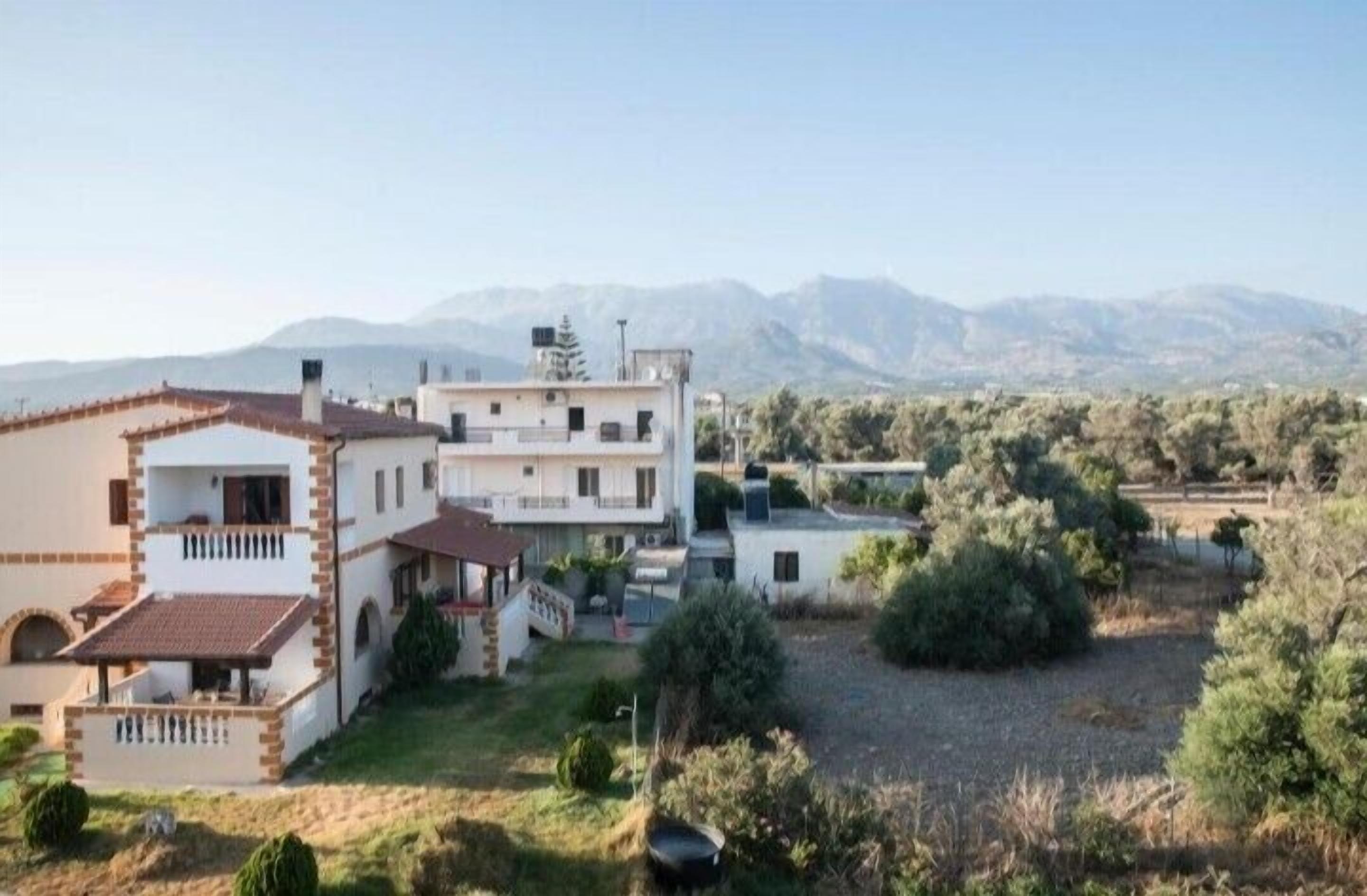 Un bâtiment à deux étages, doté d’un toit plat et d’un balcon, entouré de verdure et avec des montagnes en arrière-plan.