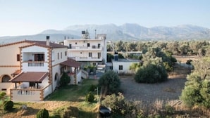 Un edificio de dos pisos con techo plano y balcón, rodeado de vegetación y montañas al fondo.