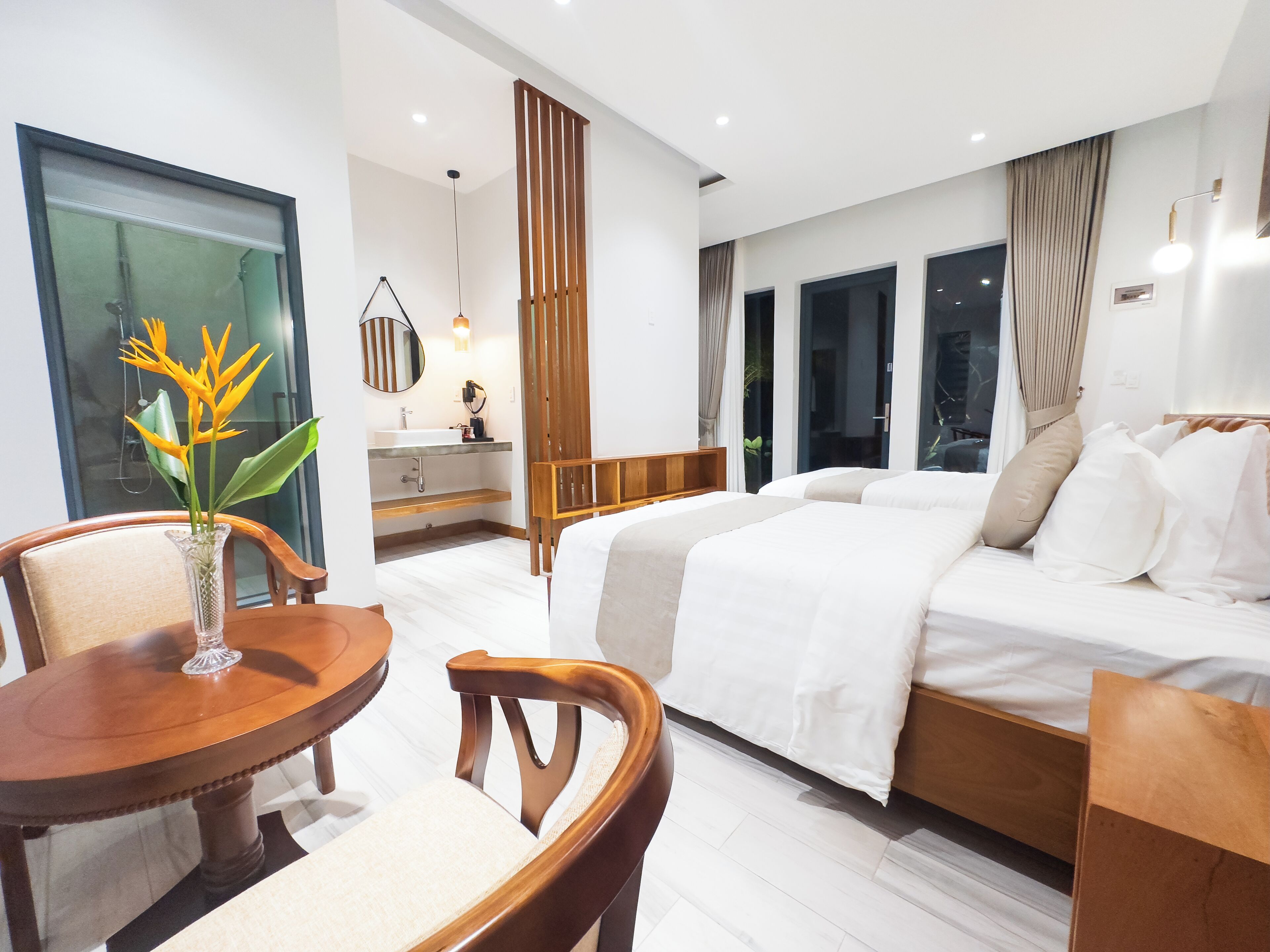 Een moderne hotelkamer met een groot bed, een zithoek met een tafel en stoelen, een badkamer met een wastafel en spiegel, en grote ramen.