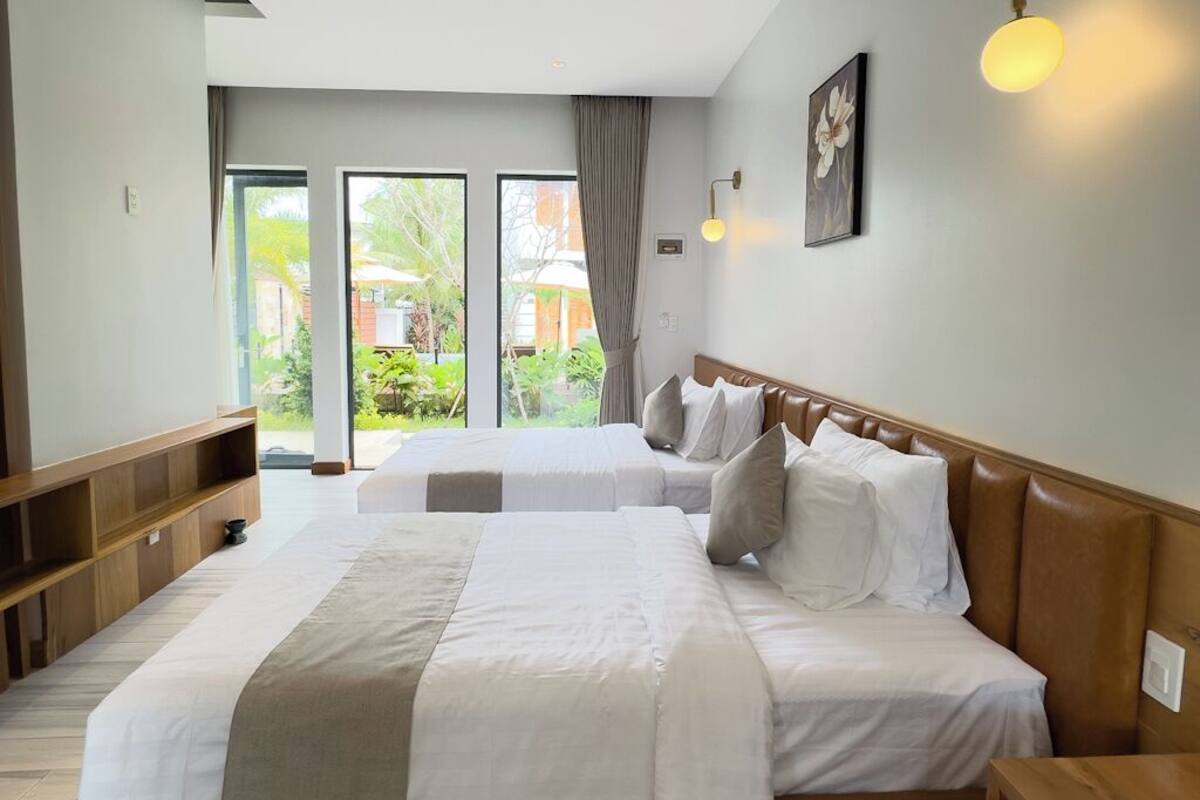 A hotel room with a large bed, a wooden headboard, a bedside table, and a view of the outside through a glass door.