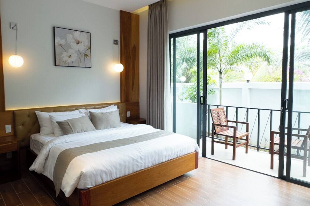 A bedroom with a large bed, a balcony with chairs, and a view of greenery.