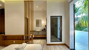 A modern hotel room with a large bed, a wooden bench, a bathroom with a large mirror, and a sliding glass door leading to a balcony with a view of palm trees. - The R&B Boutique (Kampot)
