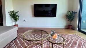 A modern living room with a flat-screen TV mounted on the wall, a grey sofa, two glass coffee tables with gold frames, and two potted plants. - FIFA World Cup - SuiteHearts Buckhead (Atlanta)