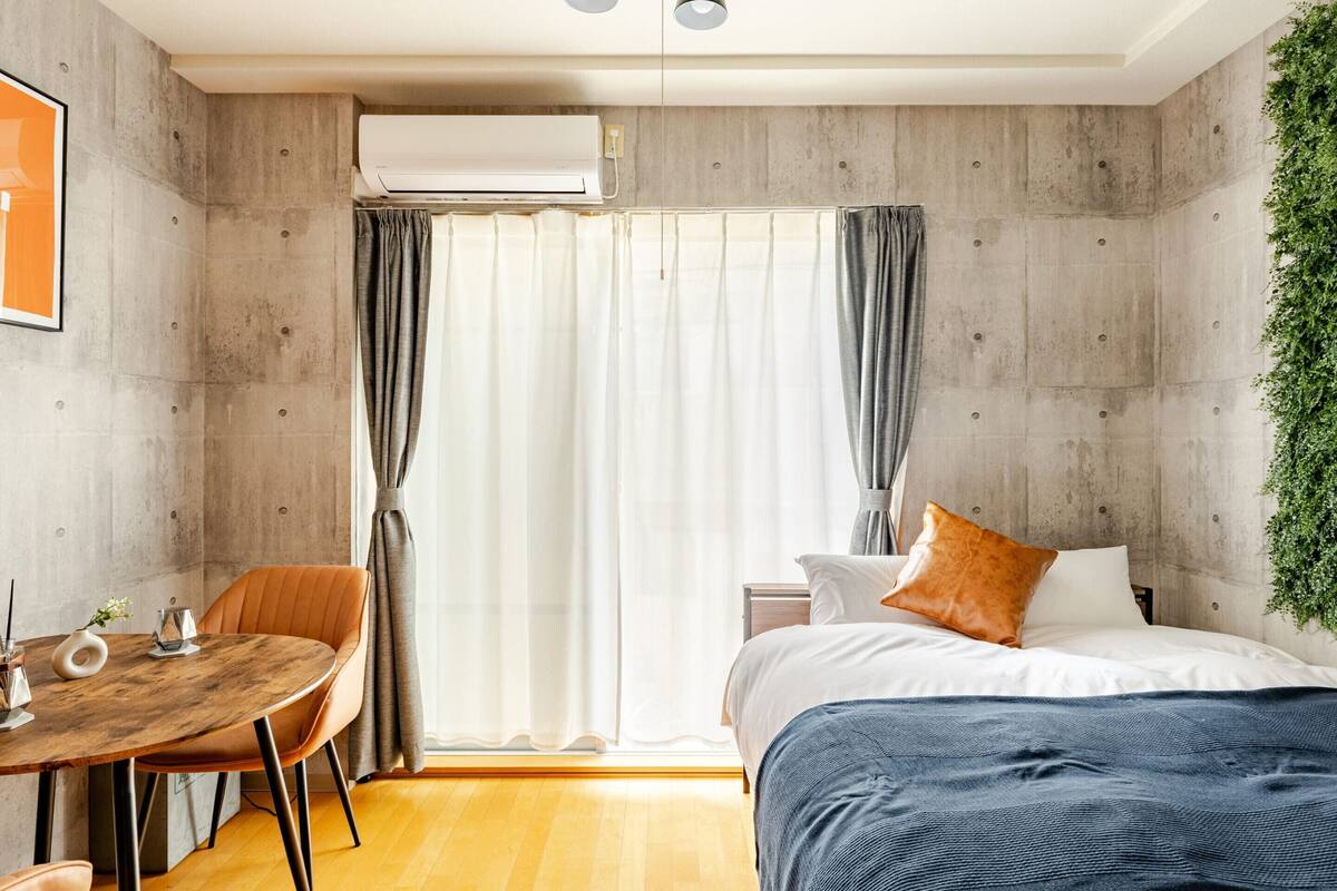 Un dormitorio con cama, mesita de noche, silla, ventana con cortinas y aire acondicionado.