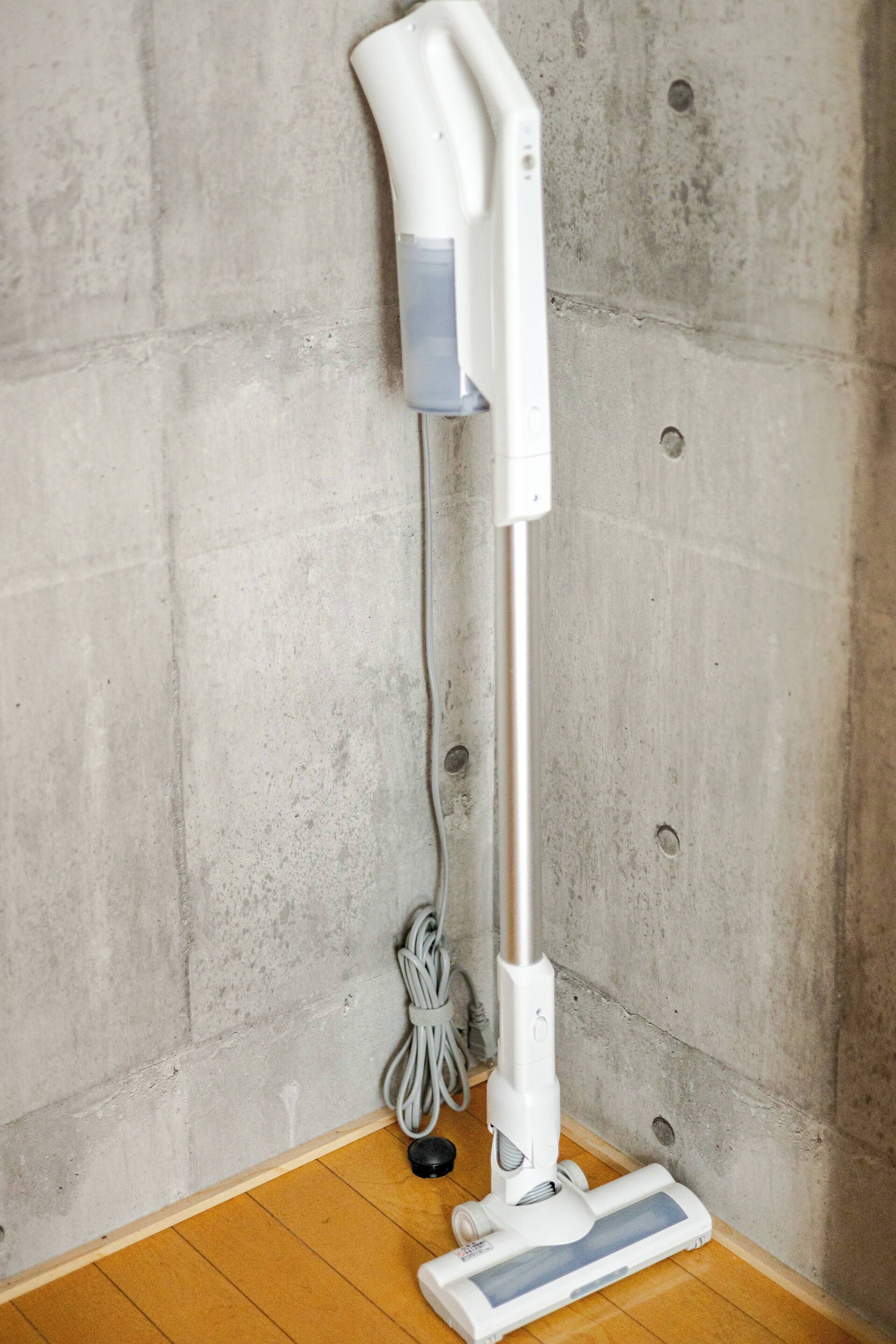 A white vacuum cleaner with a cord, standing on a wooden floor against a concrete wall.