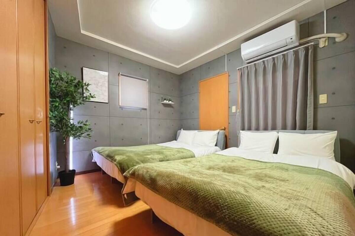 Habitación de hotel con dos camas, piso de madera, equipo de aire acondicionado, planta en maceta y cortina gris.