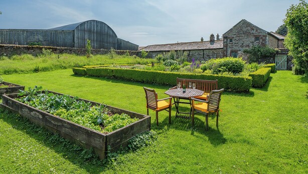 A garden with a wooden table and chairs, a stone building, and a barn in the background. - 2 bedroom accommodation in Carlisle (Carlisle)