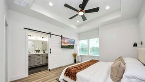 A bedroom with a bed, a ceiling fan, a TV, a sliding door to a bathroom, and a window with blinds.