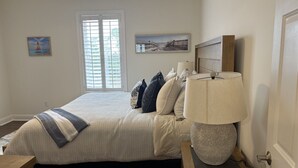 Ein Schlafzimmer mit einem Bett, einem Nachttisch mit Lampe, einem Fenster mit Jalousien und zwei Wandbildern.