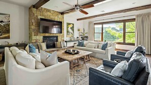 A spacious living room with a stone fireplace, a ceiling fan, and large windows offering a view of greenery. - Lake Travis Luxe Villa | 4BR | Pickleball | BBQ (Lake Travis)