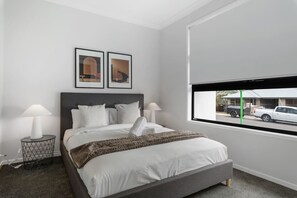 Ein modernes Schlafzimmer mit einem großen Bett, zwei Nachttischen, einem Fenster mit Blick auf parkende Autos und zwei gerahmten Bildern an der Wand.