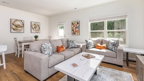 Un salon moderne avec deux canapés gris, une table basse blanche et un coin repas comprenant une table et des chaises blanches. Les murs sont ornés de tableaux abstraits et une grande fenêtre offre une vue sur la verdure.