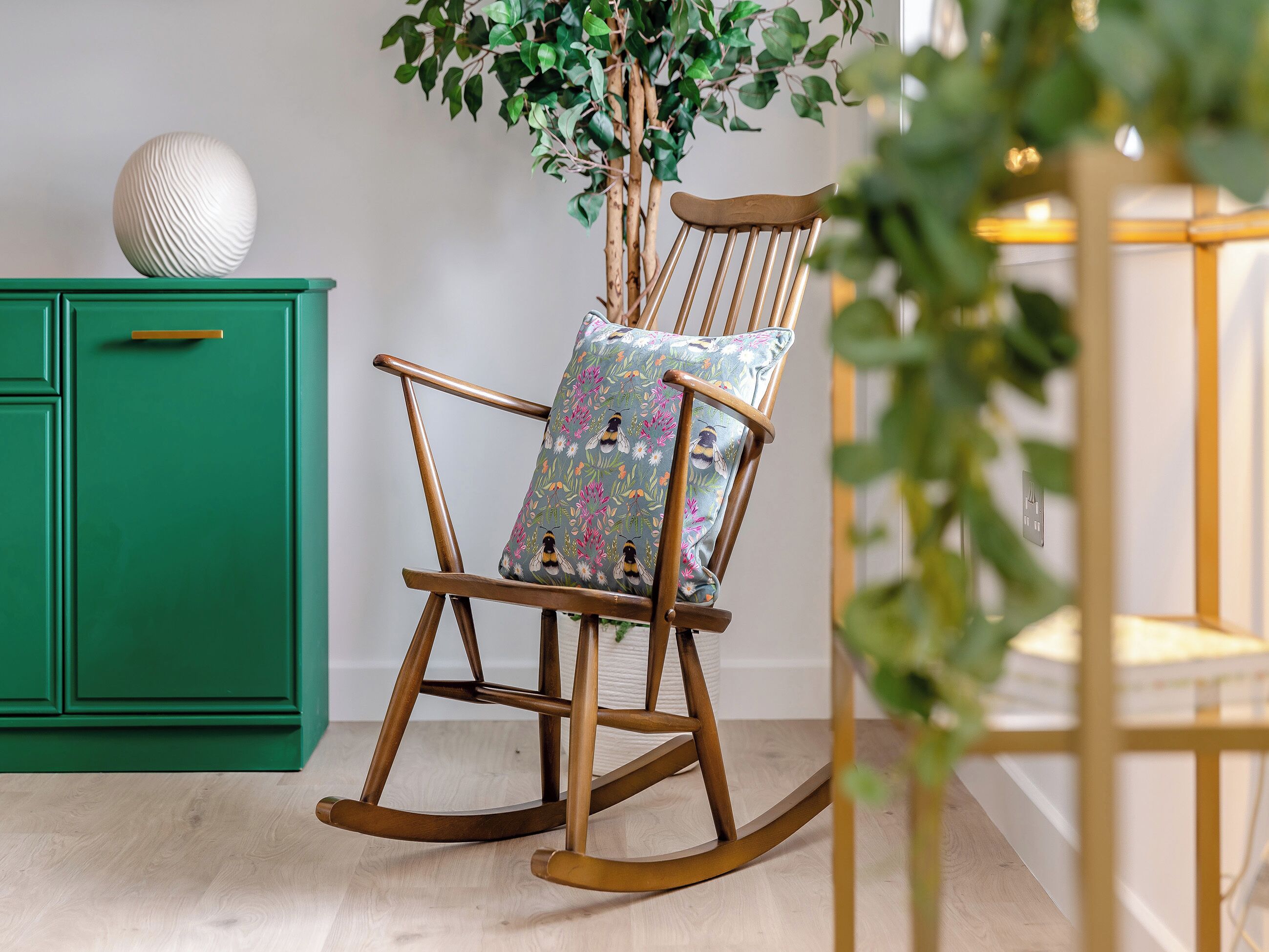 A rocking chair with a floral cushion, a green cabinet, and a plant in a room with wooden flooring.