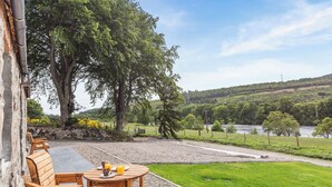 A wooden outdoor seating area with a table and chairs, overlooking a river and green landscape.