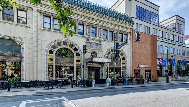 Une vue de rue d’un immeuble dont le rez-de-chaussée abrite un café et un espace de restauration, et un bâtiment en briques sur la droite, avec une enseigne indiquant « BREAKOUT ».