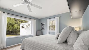 A bedroom with a large bed, two windows with curtains, a ceiling fan, and a framed picture on the wall.