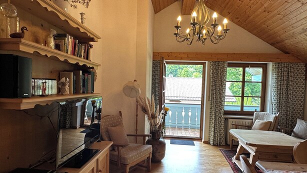 A cozy room with a wooden ceiling, a chandelier, a bookshelf, a television, a dining table, and a window with curtains. - Vacation apartment Zellerhorn 50 sqm, separate bedroom, south-facing balcony and underground garage (Aschau im Chiemgau)