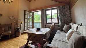 A cozy room with a wooden ceiling, a dining table with chairs, a sofa, and a large window with a view of greenery. - Vacation apartment Zellerhorn 50 sqm, separate bedroom, south-facing balcony and underground garage (Aschau im Chiemgau)