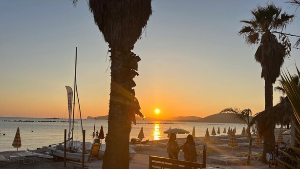 A beach scene with a sunset, palm trees, lounge chairs, and a wooden deck with a barbecue grill.