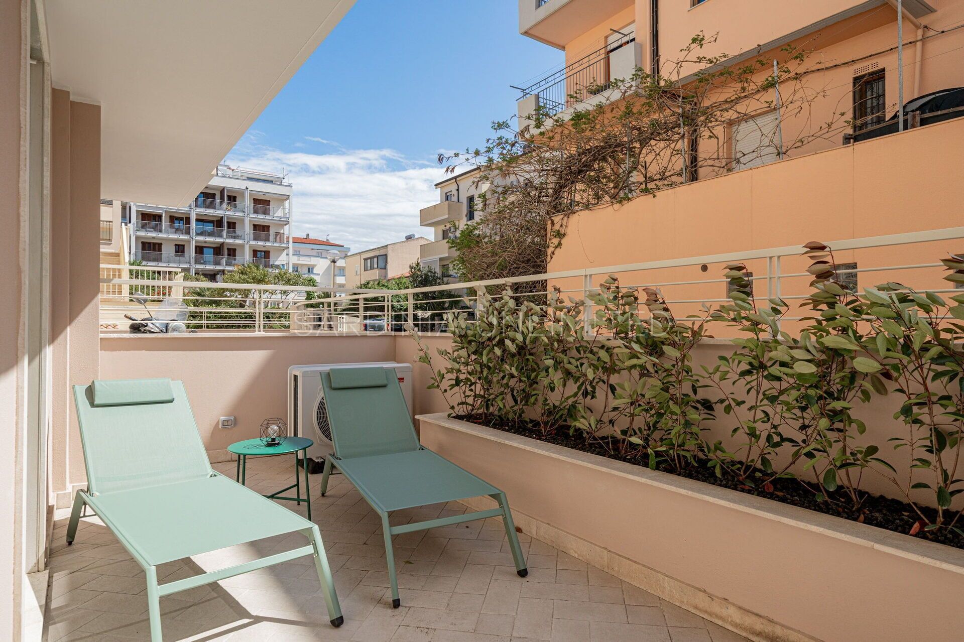 Un balcon agrémenté de deux fauteuils de jardin verts et d’une petite table, entouré d’une balustrade blanche et d’un mur bas recouvert de plantes grimpantes.