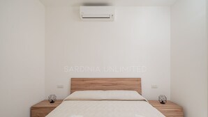 A minimalist bedroom with a wooden headboard, a bed with a textured white duvet, two bedside tables with geometric decor, and an air conditioning unit mounted on the wall.