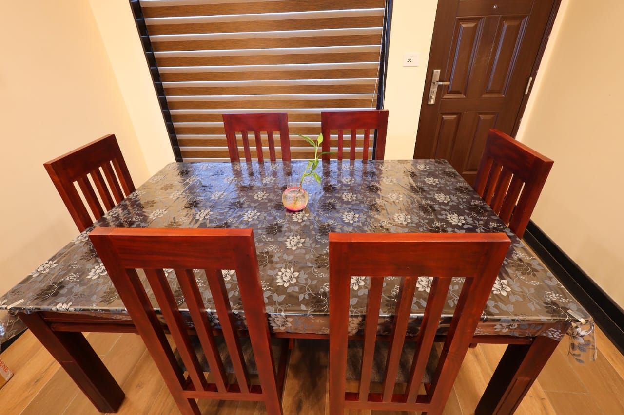 A dining table with six wooden chairs, a floral tablecloth, and a potted plant in the center.
