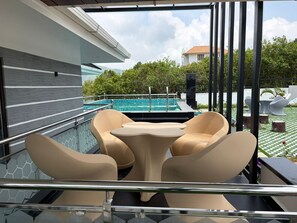 Un área de estar moderna al aire libre con barandilla de vidrio, piscina y vistas de la vegetación y edificios circundantes.