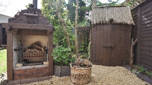 Une cheminée extérieure en briques, avec un panier de bois de chauffage devant, entourée d’une clôture en bois et d’une structure à toit de chaume.