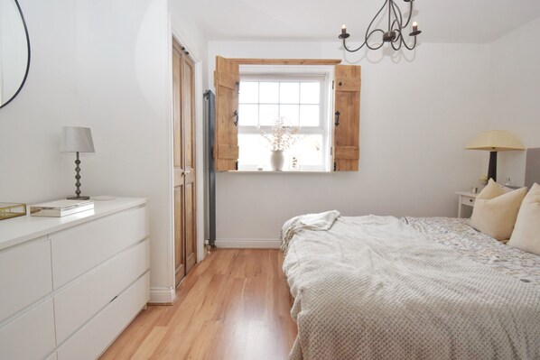 A bedroom with a wooden floor, a bed with a white blanket, a window with wooden shutters, a bedside table with a lamp, and a dresser with drawers.