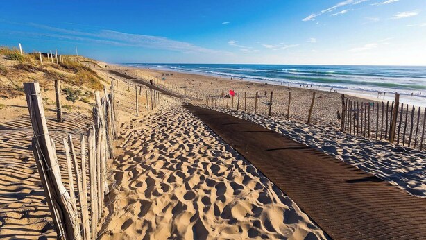 Une plage de sable avec une promenade en bois qui mène à l’océan, une clôture et un ciel bleu clair.