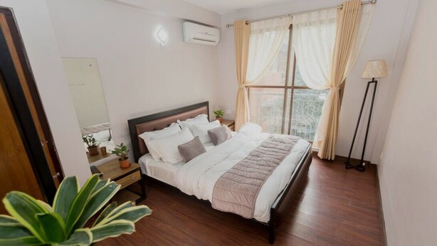A bedroom with a wooden bed, a bedside table with a lamp, a window with curtains, and a potted plant.
