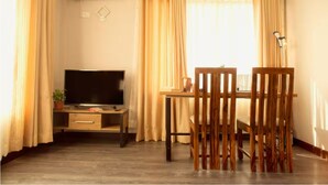 A room with a flat-screen TV on a wooden stand, a small table with a lamp, and a dining table with chairs.