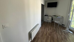 A room with a white radiator, a desk with a chair, a TV mounted on the wall, and a window with a view of greenery. - Studio 2 steps from Saint Charles station (Unknown)