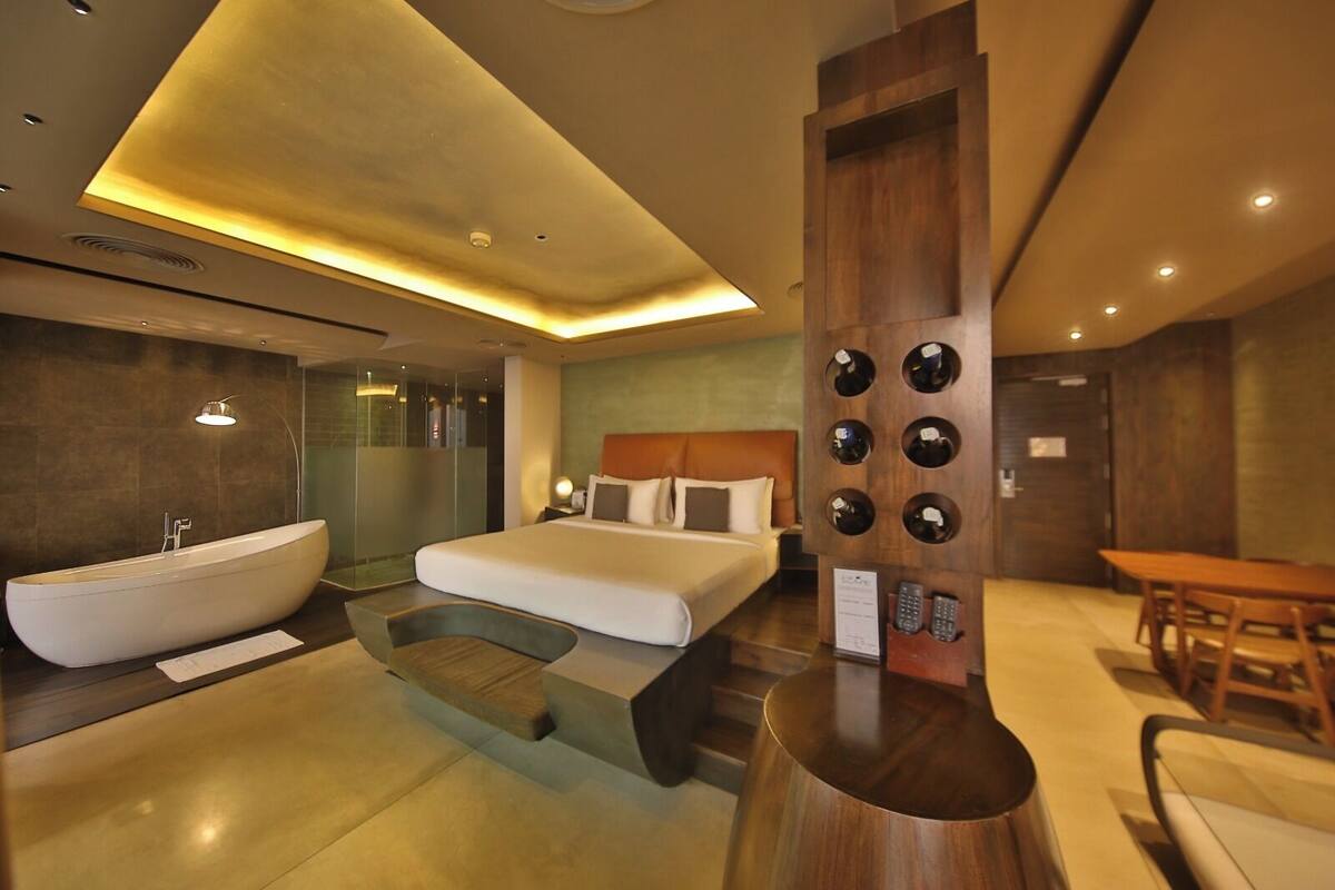 A modern hotel room with a large bed, a bathtub, a wooden table, and a unique wall-mounted wine rack.