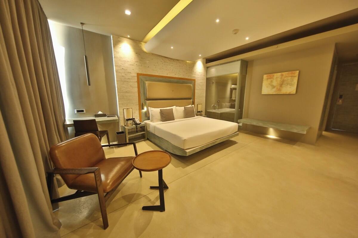 A modern hotel room with a large bed, a desk, a chair, a small table, and a bathroom with a glass shower.