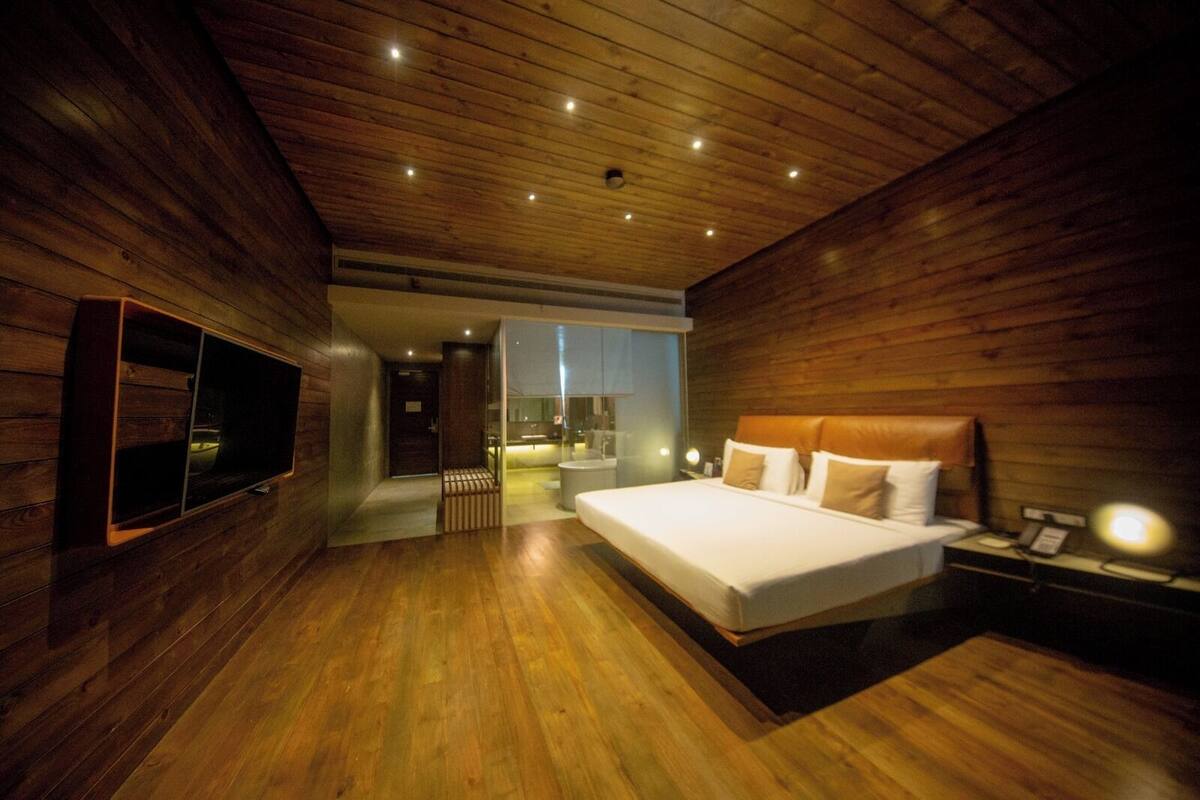 A modern bedroom with a wooden ceiling, a large bed, a TV mounted on the wall, and a bathroom visible through an open door.