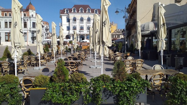 An outdoor cafe with wicker tables and chairs, surrounded by greenery and European-style buildings.