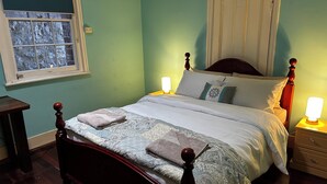 Een net opgemaakt bed met een beddekleed met een patroon en kussens, een nachtkastje met een lamp, en een raam met uitzicht op bomen.