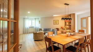 Amplia sala de estar con una mesa de comedor de madera, sillas y un sofá. Se observa una estantería llena de libros y objetos decorativos.